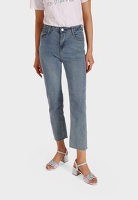 Jeans in denim chiaro con vita alta, gamba dritta e orli sfrangiati, abbinati a sandali con tacco a blocco e cinturini argentati.