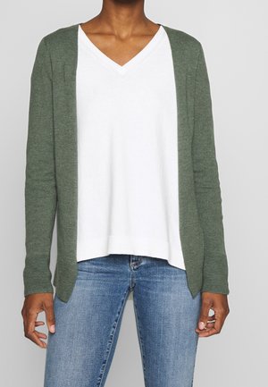 Personne portant un t-shirt blanc à col en V sous un cardigan vert olive ouvert et un jean bleu, debout devant un fond uni.