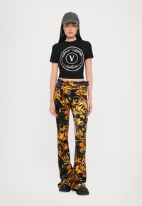 Camiseta negra corta con logo blanco, combinada con pantalones de terciopelo acampanados de patrón negro y dorado. La modelo lleva una gorra gris y zapatos negros gruesos.
