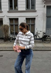 Vrouw in gestreept shirt met lange mouwen en zonnebril die een roze tas verstelt, staand op straat met gebouwen en fietsen op de achtergrond.