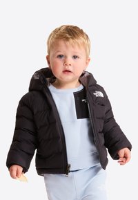 Jeune enfant aux cheveux blonds porte une doudoune noire et une tenue bleu clair, tenant un petit morceau de goûter dans la main gauche.