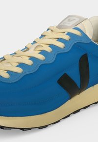 Baskets bleu et beige avec lacets crème, logo latéral noir et semelle beige avec bande de roulement noire, vues de près.