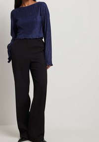 Marineblauwe geplooide blouse met lange mouwen en een gerimpelde onderrand, gecombineerd met hoge zwarte broeken. De blouse heeft een bootnek en een gestructureerde stof.