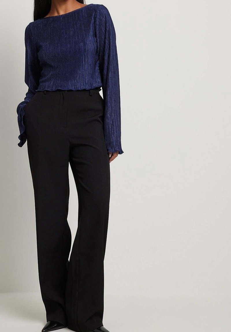 Marineblauwe geplooide blouse met lange mouwen en een gerimpelde onderrand, gecombineerd met hoge zwarte broeken. De blouse heeft een bootnek en een gestructureerde stof.