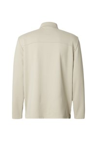 Chemise beige à manches longues avec un col à revers et une texture lisse. Caractérisée par une couture horizontale sur le haut du dos. Aucune pièce métallique visible.