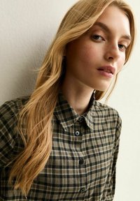 Camisa de botones a cuadros en verde y beige con cuello, que presenta una textura suave y botones negros. Cabello largo y ondulado en el fondo.