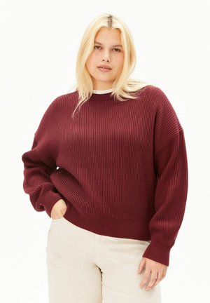 Jeune femme aux cheveux blonds portant un pull côtelé bordeaux et un pantalon beige clair, posant avec une main dans la poche.