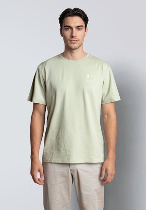 Mann mit dunklem Haar, der ein hellgrünes T-Shirt mit kleinem weißem Smiley und hellbeige Hosen trägt, vor einem einfarbigen Hintergrund stehend.