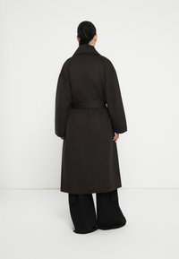 Abrigo largo de color marrón oscuro con un cuello ancho y un cinturón. Presenta mangas sueltas y una silueta recta. Llevado sobre pantalones de pierna ancha negros.