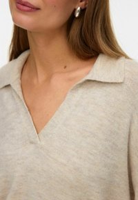 Beżowy sweter z dekoltem w serek, wykonany z miękkiego materiału dzianinowego z kołnierzykiem. Posiada delikatną teksturę i nie ma wyraźnych wzorów ani ozdób.