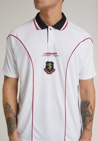 Hvid polo shirt med røde accenter og sort krave; prydet med "UMBRO Football League" logo og emblem. Korte ærmer med broderede detaljer.