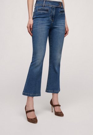 Blue denim flared jeans with a mid-rise waist, two front pockets, two buttons, and frayed hems. Paired with brown high-heeled shoes.