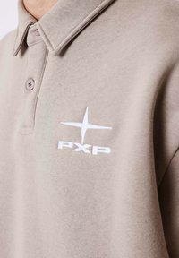 Polo beige in tessuto morbido con colletto e due bottoni. Presenta un logo ricamato bianco "PXP" e un dettaglio a forma di stella sul petto.