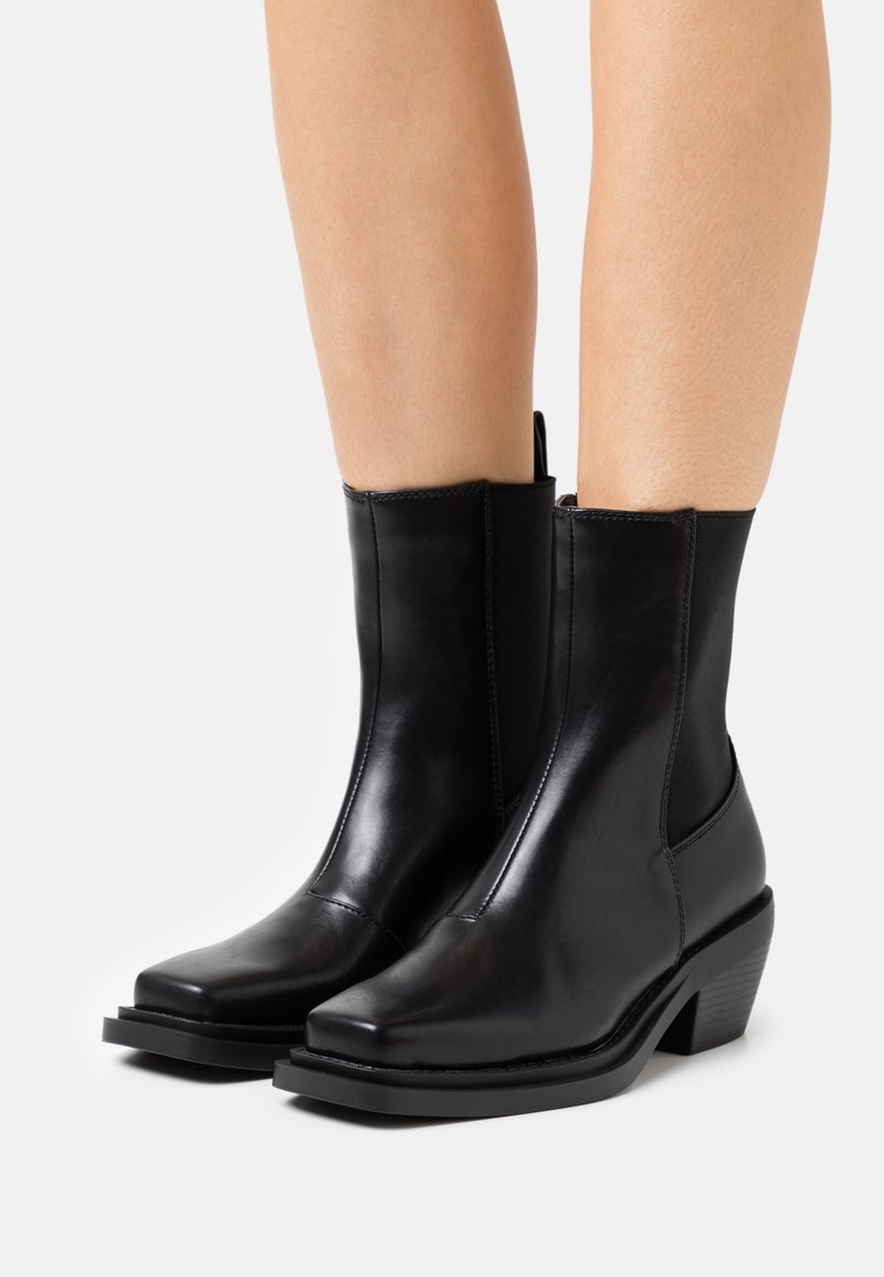 Bottes courtes en cuir noir avec un bout carré, des panneaux latéraux élastiques et un talon bas empilé. Texture lisse et détails de couture minimaux.