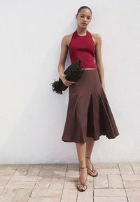 Punainen halterneck-toppi, joka on yhdistetty ruskeaan levenevään hameeseen, jossa on poimutuksia. Malli pitää käsissään mustaa, teksturoitua clutch-laukkua, jossa on fringet. Vaalea tausta.