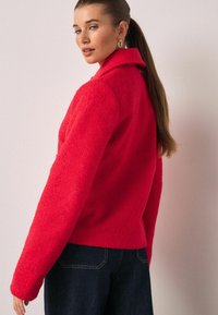 Rød fleecejakke med krave, lange ærmer og en tekstureret finish. Kombineret med mørke denimjeans og et diskret tilbehør.