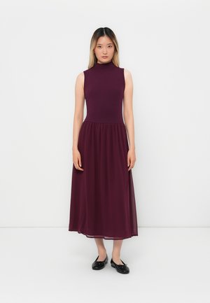 Mauve ermeløs kjole med tettsittende liv og flytende ankel-langt skjørt, laget av lett stoff. Svarte leiligheter fullfører antrekket.