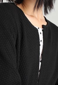 Svart texturerad jacka med rundad halsringning, med ett vadderat tyg och öppen front, tillsammans med en vit skjorta prydd med svarta prickar.