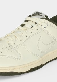 Λευκό και ελιά αθλητικό παπούτσι Nike με κορδόνια, κεντημένο λογότυπο στο πλάι, διάτρητα στο μπροστινό μέρος και ετικέτα με λογότυπο στη γλώσσα.