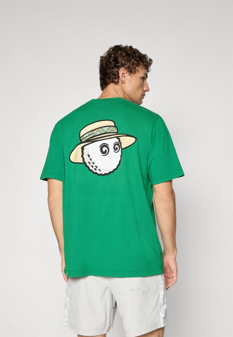 Mann som har på seg en grønn t-skjorte med et grafisk bilde av en golfball med spiraløyne og en stråhatt, sammen med lyse shorts.