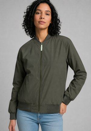 Frau mit lockigem Haar, die eine grüne Bomberjacke mit Reißverschluss und hellblaue Jeans trägt, steht mit einer Hand in der Tasche vor grauem Hintergrund.