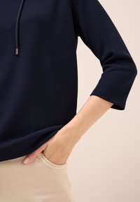 Top a righe blu navy con scollatura rotonda e maniche a tre quarti, abbinato a pantaloni beige rilassati. Mano appoggiata nella tasca laterale.