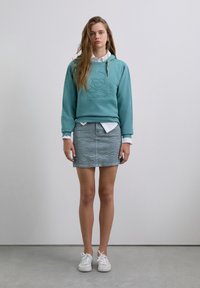 Young woman wearing teal hoodie with skull logo, white shirt, gray patterned mini skirt, and white sneakers standing against plain background.