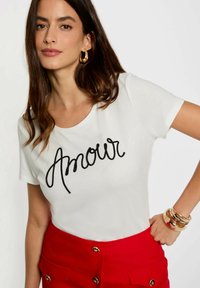 Witte katoenen t-shirt met zwart geborduurd tekst "Amour." Gecombineerd met een rode rok, met gouden hardware accenten. Soepele textuur.