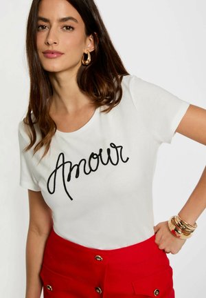 T-shirt en coton blanc avec le texte noir brodé "Amour". Associé à une jupe rouge, mettant en valeur des accents en métal doré. Texture lisse.