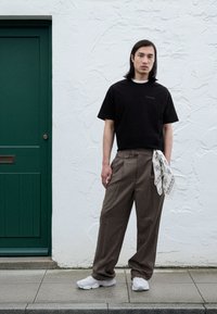 Svart t-skjorte med korte ermer og subtil merkevare, brune bukser med tynne striper, et hvitt mønstret skjerf og hvite joggesko foran en strukturert hvit vegg.