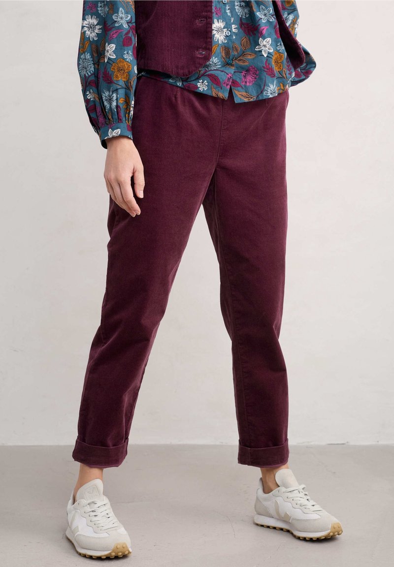 Pantalons en velours côtelé bordeaux avec des jambes fuselées, présentant un ourlet retourné. Assortis à des baskets blanches. La tenue est complétée par un chemisier floral.