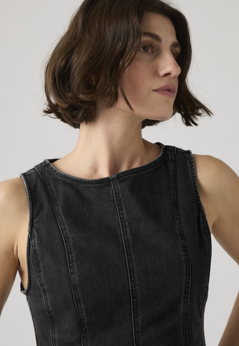Young woman with short dark hair wearing a sleeveless black denim top, looking slightly to her right against a plain light background.