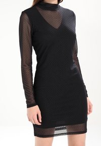 Schwarzes Kleid mit langen, durchsichtigen, gepunkteten Ärmeln und einem tiefen V-Ausschnitt. Glatte Textur, figurbetontes Design, knielang mit einem semi-transparenten Rock.