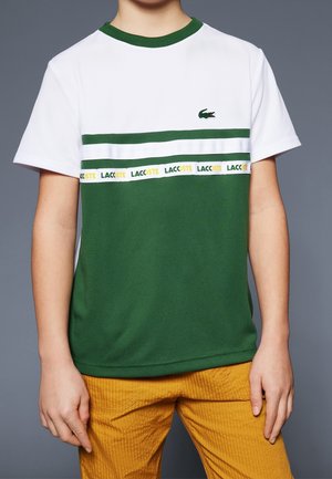Barn iført en hvid og grøn Lacoste t-shirt med logo og gule bukser, stående mod en grå baggrund.