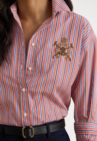 Sarkana, zilā un baltā krāsā svītraina pogas aizdares krekls ar izšūtu zelta ģerboni, klasisku apkakli un aproču piedurknēm.