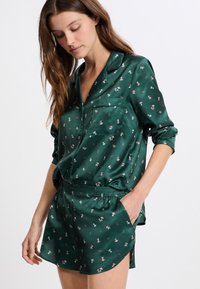 Grünes Satin-Pyjama-Set mit floralem Muster; langärmlige, knöpfbare Bluse und Shorts mit elastischem Bund. Weiche Textur und glatter Glanz.