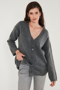 Naine, kes kannab tumedat halli nööbitavat kudumit, heledaid halli teksaseid, hõbedasi rõngakujulisi kõrvarõngaid ja käevõrusid, poseerib ühevärvilise heleda tausta ees.