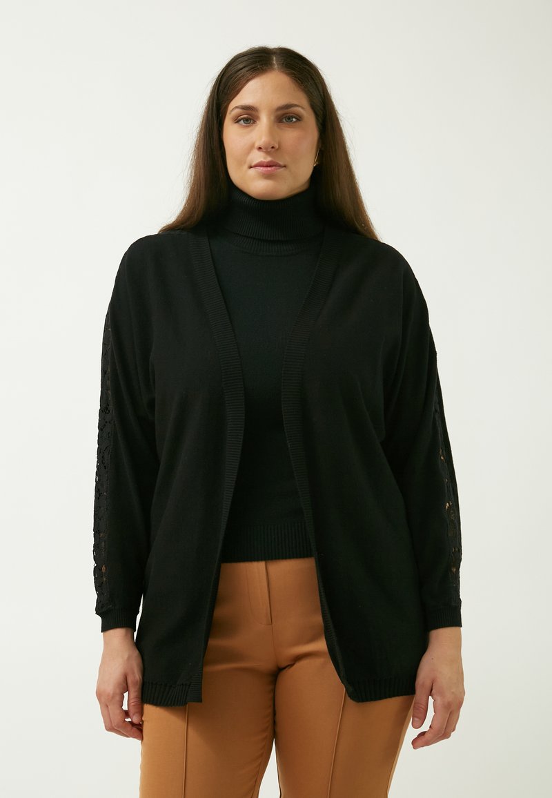 Sort cardigan med åben front, prydet med blondeaccenter på ærmerne. Stoffet er glat og strikket, lagd over en sort polo.