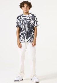 Schwarzes und weißes grafisches T-Shirt mit kurzen Ärmeln und Palmblattmuster, kombiniert mit weißen Hosen und weißen Sneakers. Einfaches Design und lockere Passform.