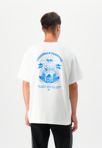 White cotton t-shirt with oversized fit, featuring a blue graphic design on the back, including festival themes and city names.