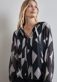 Fließende schwarz-weiße Bluse mit geometrischem Rautenmuster, langen transparenten Ärmeln und einem V-Ausschnitt mit Knopfverschluss.