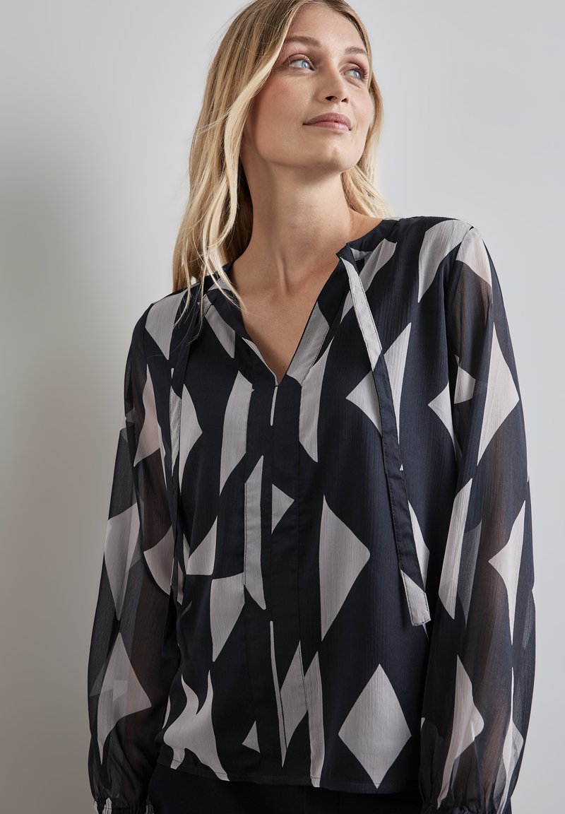 Fließende schwarz-weiße Bluse mit geometrischem Rautenmuster, langen transparenten Ärmeln und einem V-Ausschnitt mit Knopfverschluss.