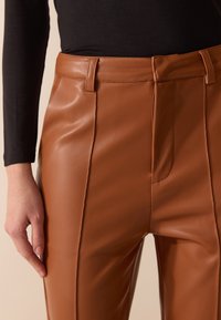 Brune faux skinnbukser med en glatt tekstur, rett passform og frontfolder. Har flatt liv og sidelommer.