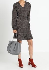 Robe portefeuille noire à motifs floraux avec manches longues, ourlet volanté, associée à un sac seau en cuir gris et des bottines à talons en suede rouge.