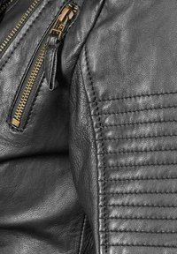Detail van een zwart leren jack met gouden ritssluitingen, een textuurafwerking en een verticale stiknaadontwerp langs de schouder en mouw.