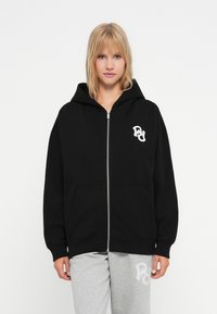 Čierna mikina na zips s priestranným strihom, klokaními vreckami a bielym logom na ľavej hrudi. Tkanina z bavlny s jemnou textúrou.