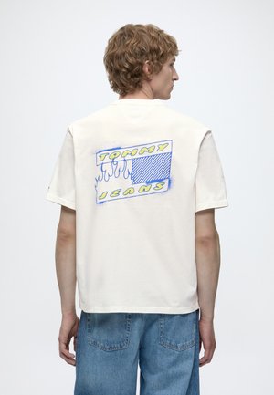 Młody mężczyzna z kręconymi włosami, ubrany w białą koszulkę Tommy Jeans z niebiesko-żółtą grafiką płomieni na plecach, w połączeniu z niebieskimi dżinsami.