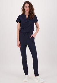 Marineblauwe jumpsuit met korte mouwen, een v-hals, een trekkoord in de taille en zijzakken, gecombineerd met witte sneakers. Glad stofoppervlak.