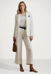 Mulher com blazer branco, blusa estampada, calças largas de cintura alta, sandálias de tiras castanhas, e segurando uma mala castanha.