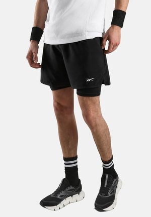 Man draagt zwarte Reebok shorts met ingebouwde compressielaag, zwart-witte sneakers, zwarte polsbandjes en gestreepte zwarte sokken.